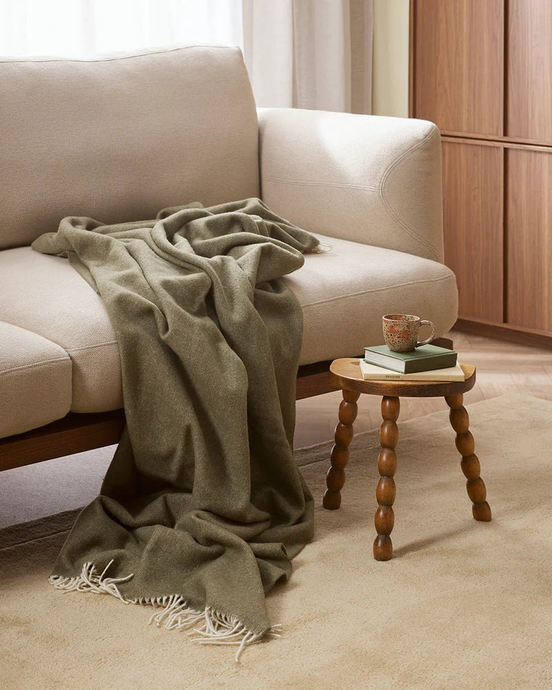 Green blanket draped over a beige sofa with a wooden side table and books in the background.