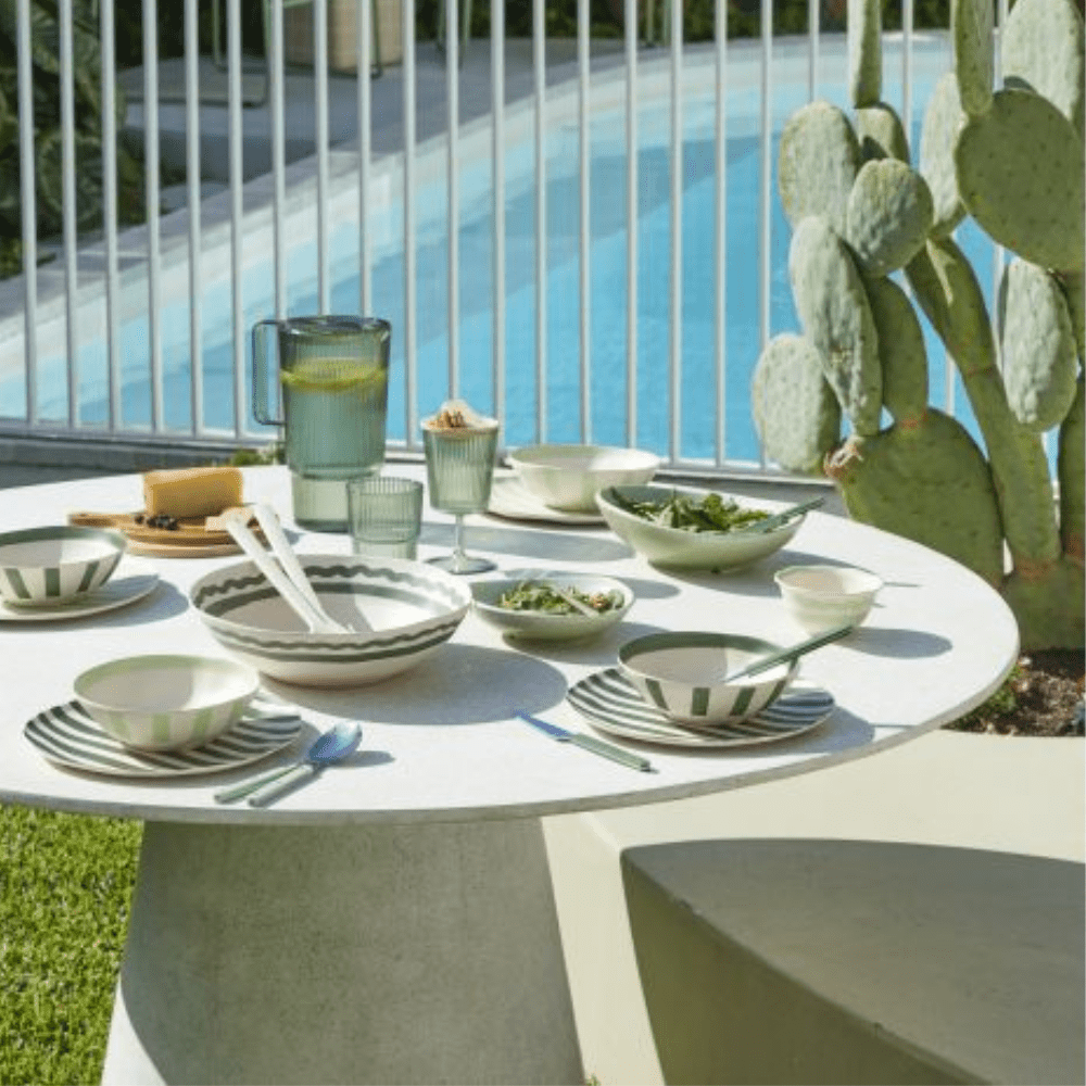 Outdoor dining setup with a table by a poolside, featuring green and white ceramic dishes and a cactus plant.