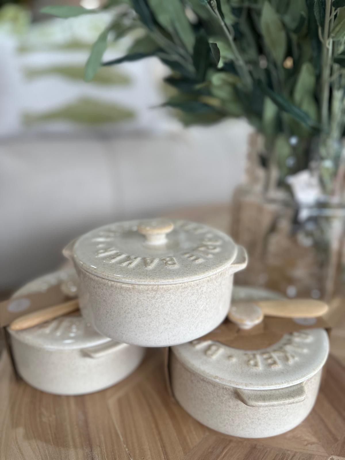 Set of three ceramic brie bakers with lids on a wooden surface, with a blurred background of greenery.